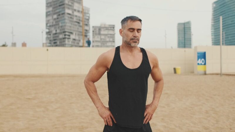 Muscular older man standing on the beach in a black tank top, appearing thoughtful post-exercise