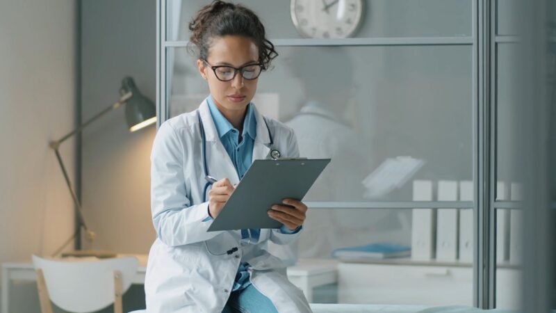 Young female doctor writing notes on a clipboard in a hospital office, wearing a white coat and stethoscope