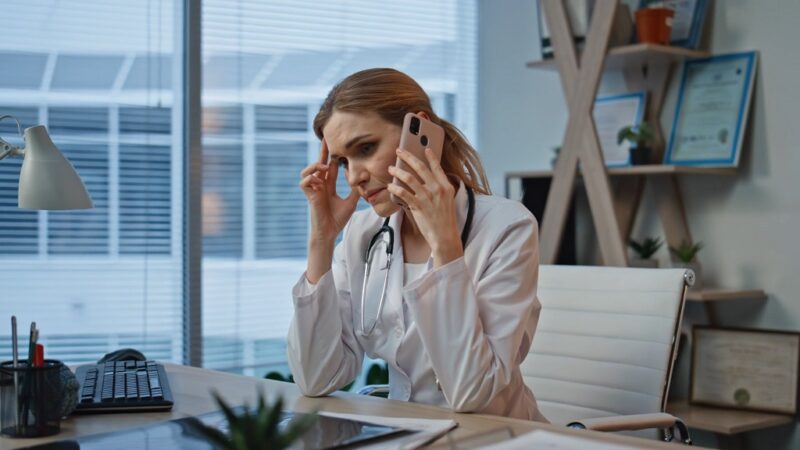 Female doctor in a hospital office looking stressed while talking on the phone, holding her head with one hand