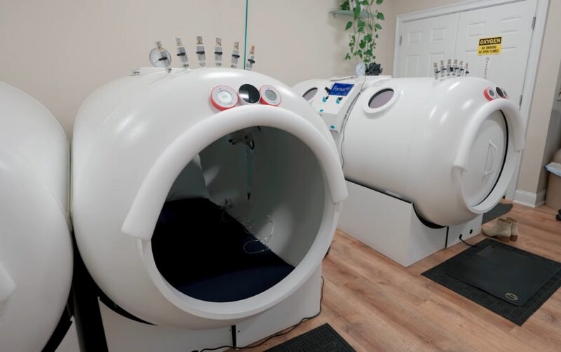 Hyperbaric oxygen therapy chambers in a clinical treatment room, featuring white pressurized units with gauges and patient seating areas